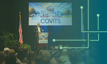 CIO of the Commonwealth Bob Osmond addresses a crowd from behind a podium on stage with 'COVITS 2025' imagery on the wall behind him and on the podium.